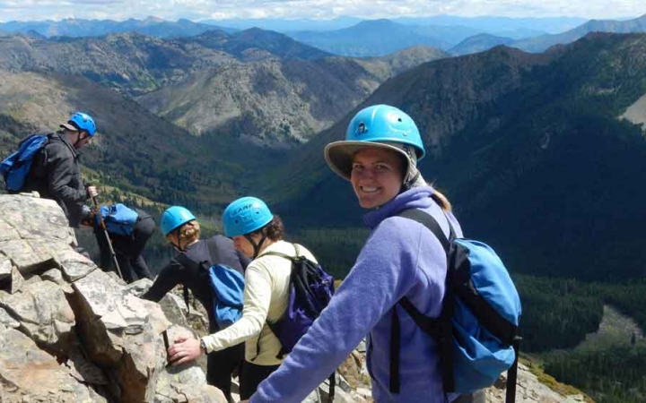 a group of students wearing helmets traverse a rocky landscape with mountains in the background